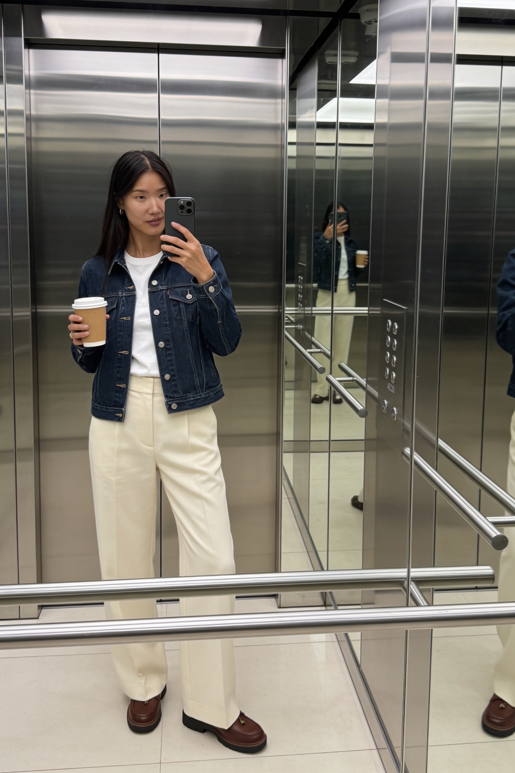 Woman taking an elevator mirror selfie wearing a dark Levi's denim trucker jacket, wide cream tailored trousers, and brown leather Clarks loafers with a takeaway coffee.